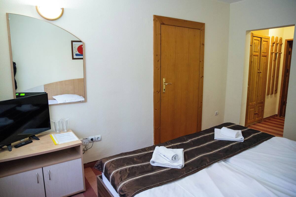 A hotel room with a bed, a desk with a computer and glasses, a mirror, and a door.