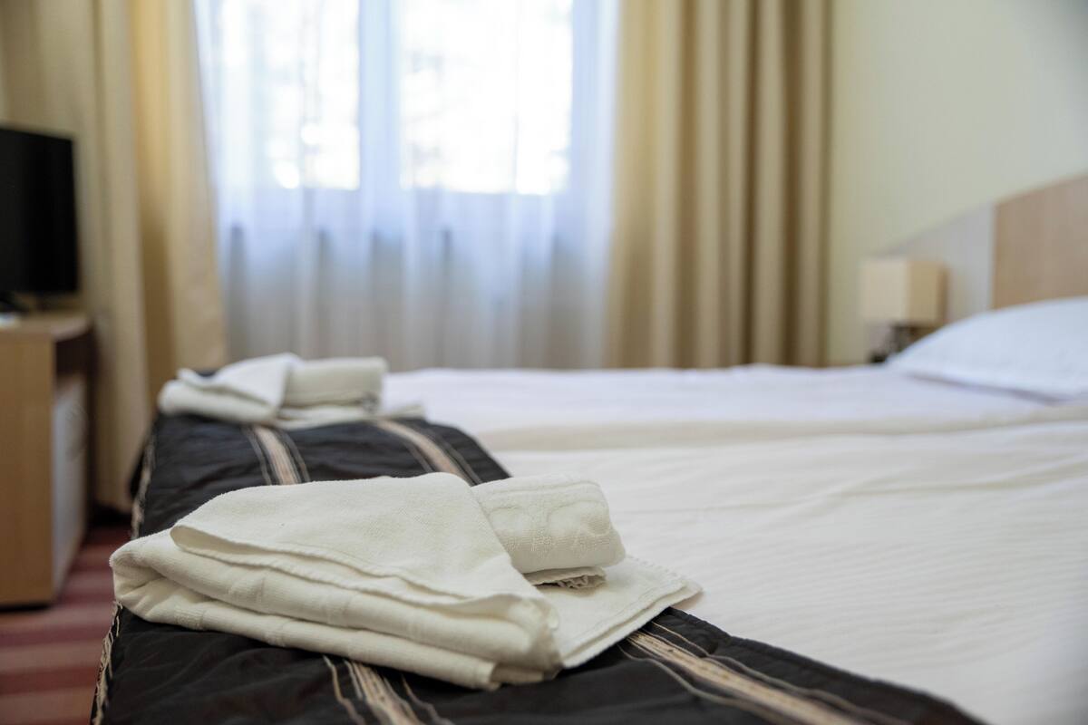 A hotel room with a neatly made bed, a television on a stand, and a window with curtains.