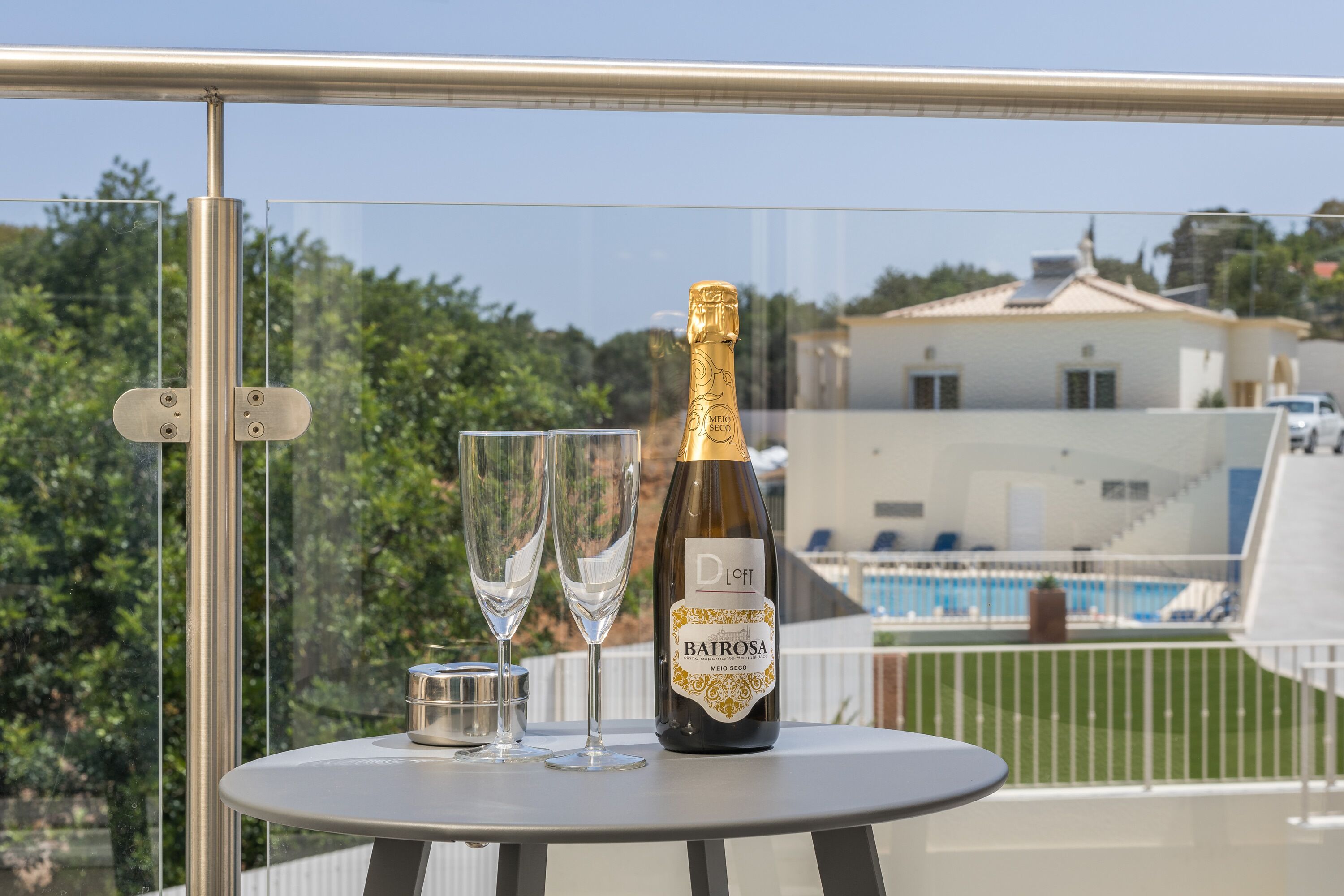 A bottle of Bairrosa champagne, two glasses, and a small container on a balcony table with a pool view.