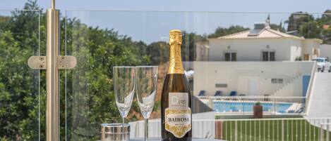 A bottle of Bairrosa champagne, two glasses, and a small container on a balcony table with a pool view.