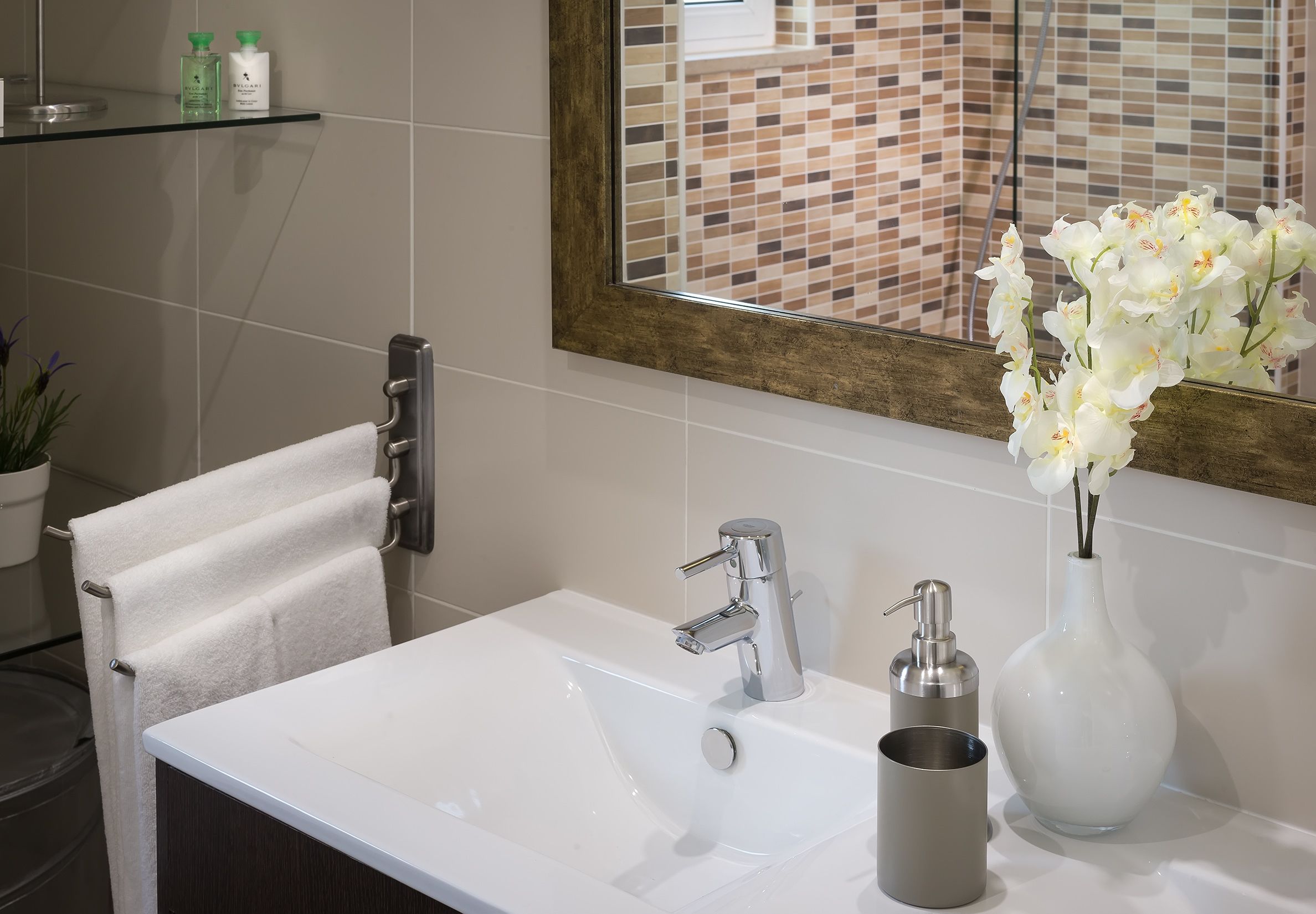 Ein Badezimmer mit einem weißen Waschbecken, einem chromfarbenen Wasserhahn und einem Spiegel darüber. Ein Handtuchhalter mit einem weißen Handtuch und eine Vase mit weißen Blumen stehen auf der Waschtischablage.