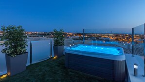 Una terraza en la azotea con jacuzzi, plantas en macetas y vista a la ciudad al anochecer.