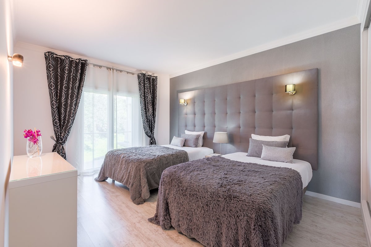 A hotel room with two beds, a large padded headboard, bedside lamps, and a desk with a vase of flowers.