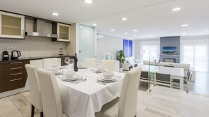 Un comedor moderno con una mesa blanca dispuesta para cuatro personas, una cocina con estufa integrada y una sala de estar con sofá y televisor.