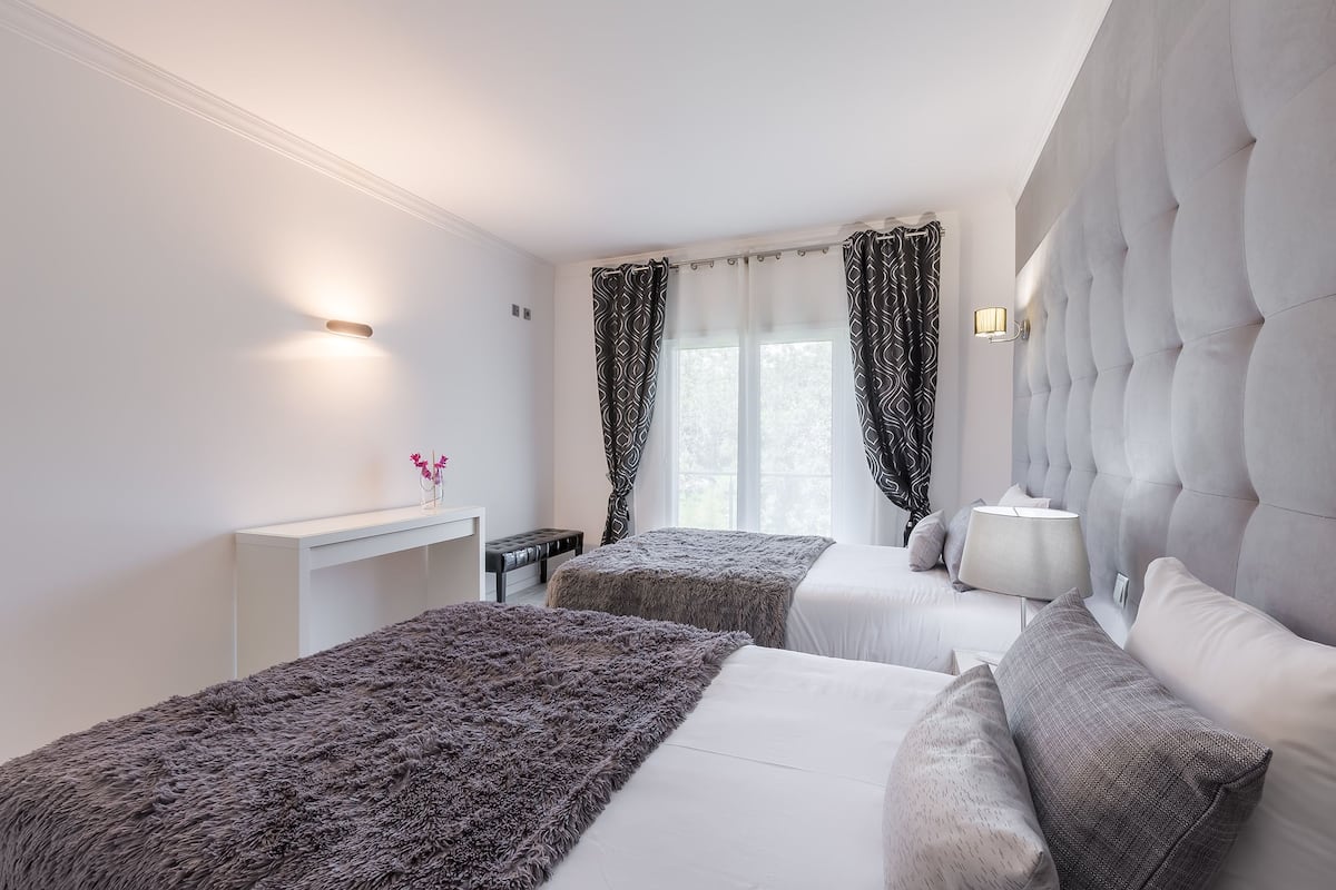 A modern hotel room with two beds, a tufted headboard, a bedside table with a lamp, and a window with patterned curtains.