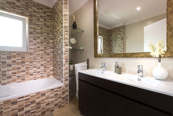 A modern bathroom with a tiled wall, a large mirror, a white sink, and a bathtub.