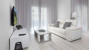 Una sala de estar moderna con un sofá blanco, una mesa de centro, un televisor sobre un mueble bajo y cortinas translúcidas.