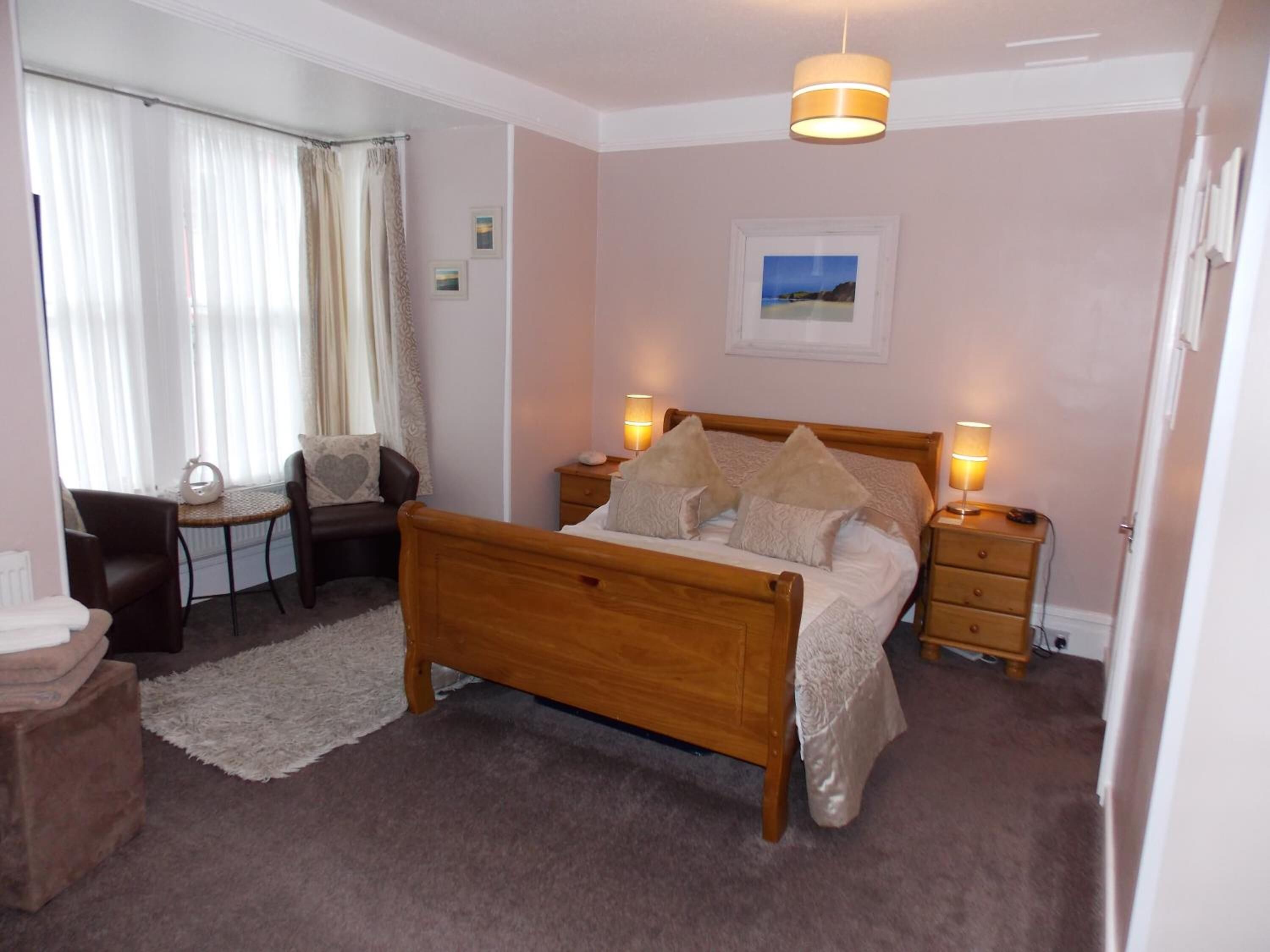 A bedroom with a wooden bed, bedside tables, a chair, a small table, and a window with curtains.