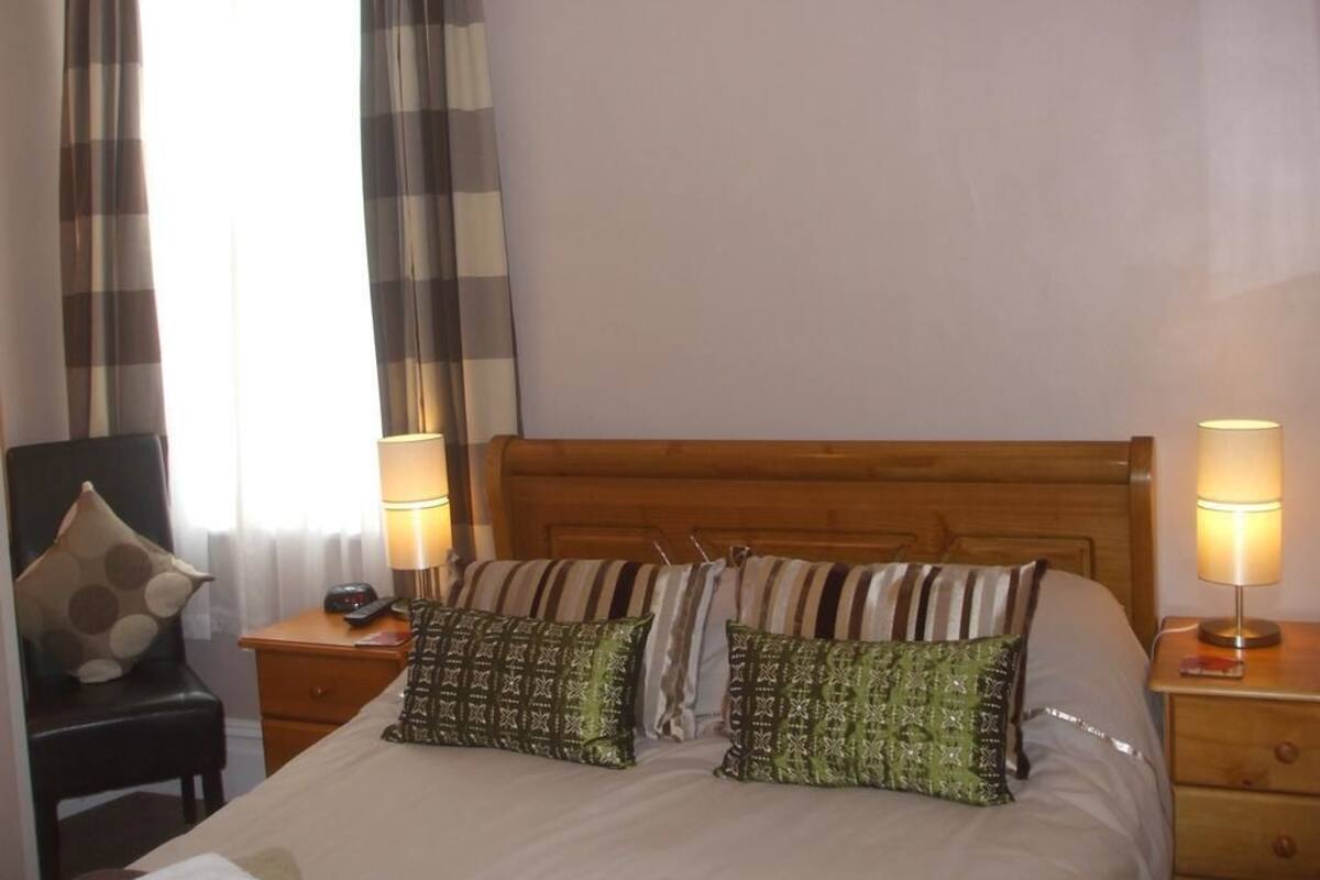 A hotel room with a wooden headboard, two bedside lamps, a chair with a pillow, and a bed with patterned pillows.