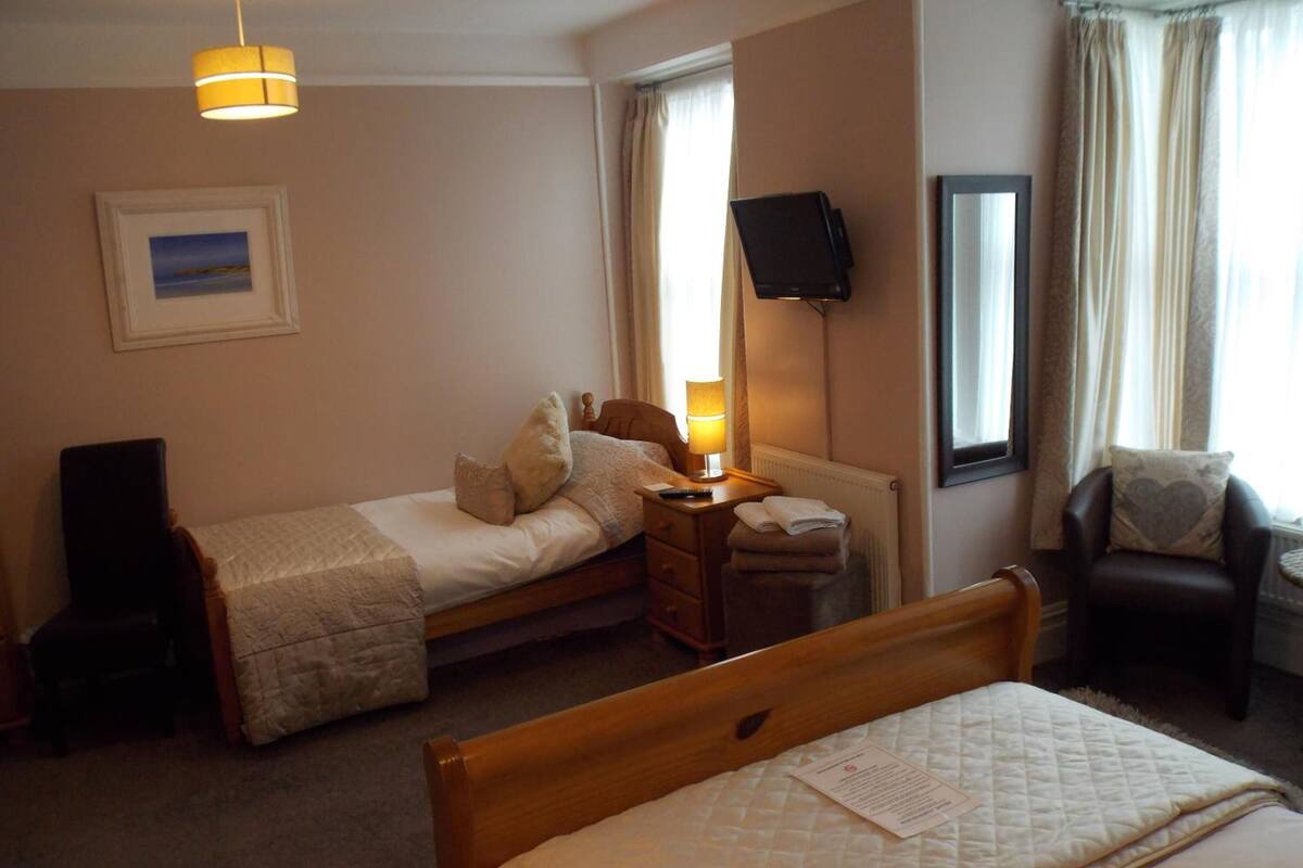 A hotel room with a single bed, a smaller bed, a desk with a lamp, a chair, a TV mounted on the wall, and a framed picture on the wall.