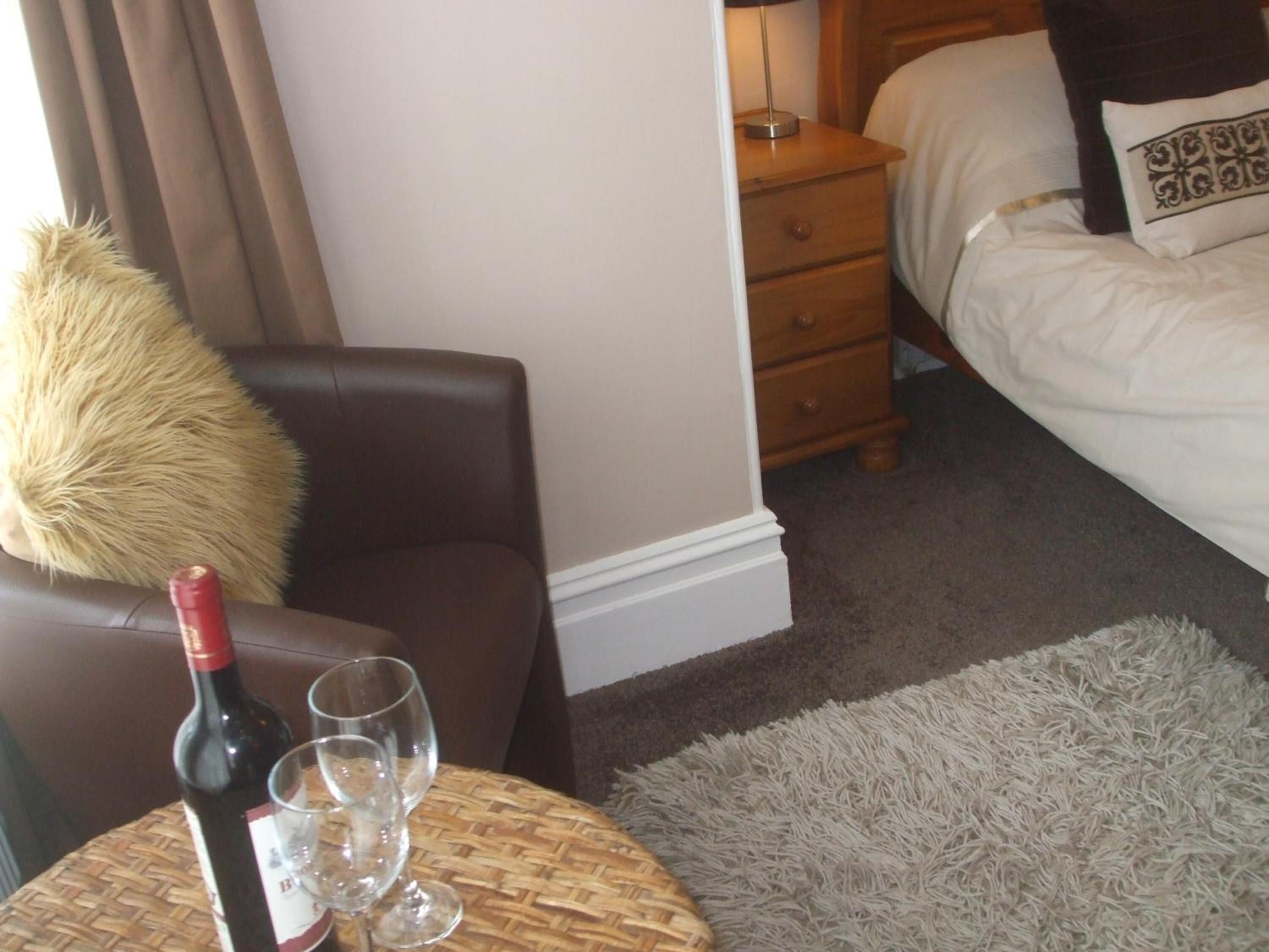 Una habitación de hotel con una cama, una mesita de noche, una silla, una pequeña mesa redonda con una botella de vino y copas, y una alfombra mullida.