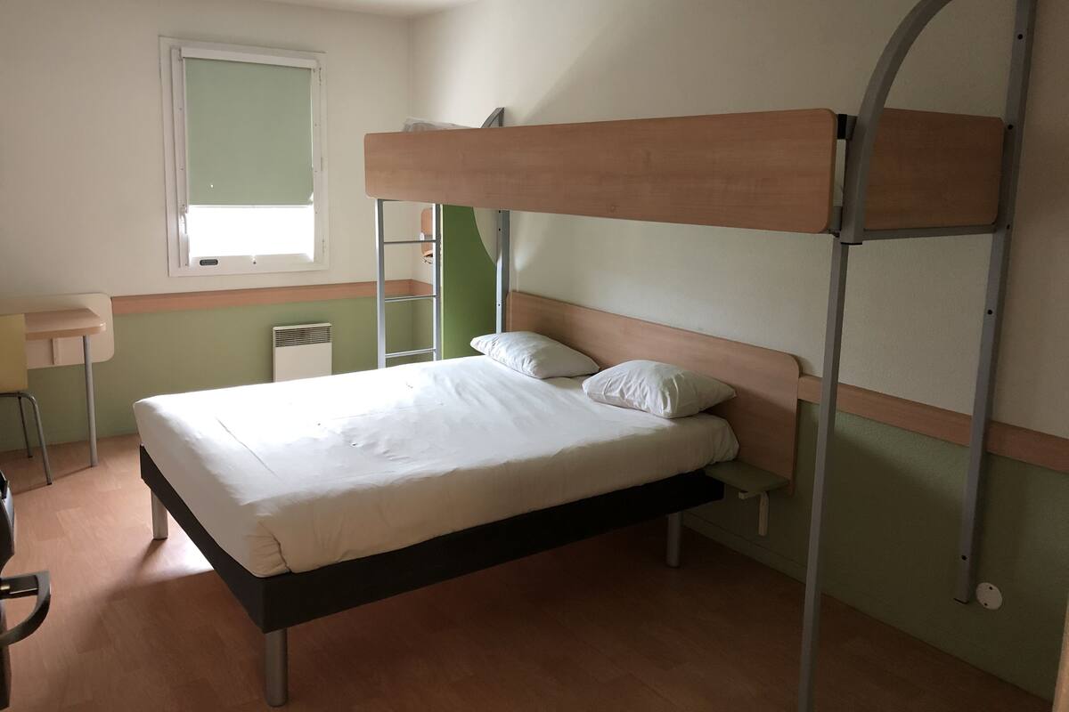 Ein Etagenbett mit Holzkopfteil, einem Bett unten, einem kleinen Schreibtisch und einem Fenster.