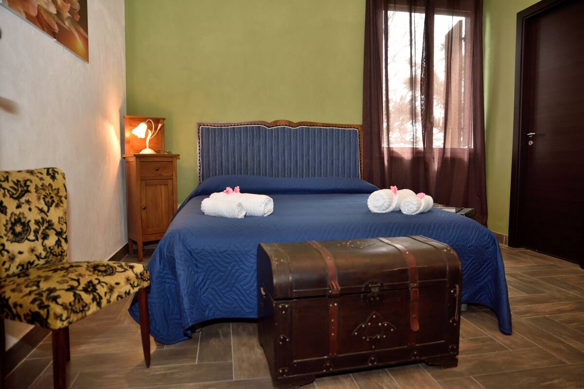 Ein ordentlich gemachtes Bett mit blauer Bettdecke, zwei zusammengerollten Handtüchern und einem hölzernen Kopfteil. Ein Nachttisch aus Holz mit Lampe und kleinem Schrank steht neben dem Bett. Ein alter Koffer liegt auf dem Boden neben dem Bett.
