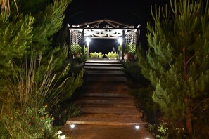 Un chemin illuminé qui mène à une pergola en bois, elle-même entourée de verdure.