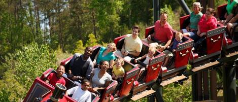 A group of people on a wooden roller coaster with red cars, surrounded by greenery.