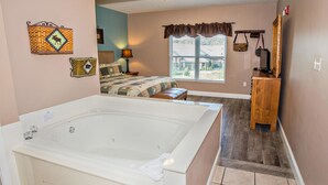 A bedroom with a large bathtub, a bed with a patterned bedspread, a window with a view, and a wooden dresser. - Bear Crossing 202 (Pigeon Forge)