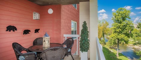 A balcony with wicker chairs and a table, a red wall with bear decorations, a small window, and a view of a river and trees.