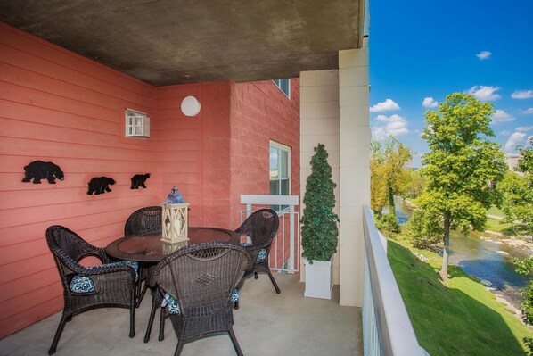 A balcony with wicker chairs and a table, a red wall with bear decorations, a small window, and a view of a river and trees.