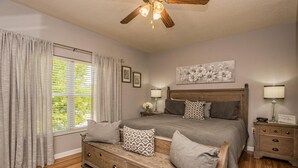 Un dormitorio con una cama, mesitas de noche, un banco de madera, un ventilador de techo y una ventana con cortinas.