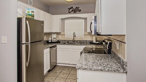Una cocina con gabinetes blancos, un refrigerador de acero inoxidable, una encimera de granito y una ventana con persianas.