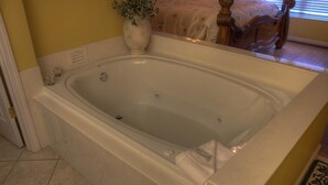 Eine große, rechteckige Badewanne mit Holzrahmen, ein kleiner Vasen mit einer Pflanze und ein Bett mit Holz Kopfteil im Hintergrund.