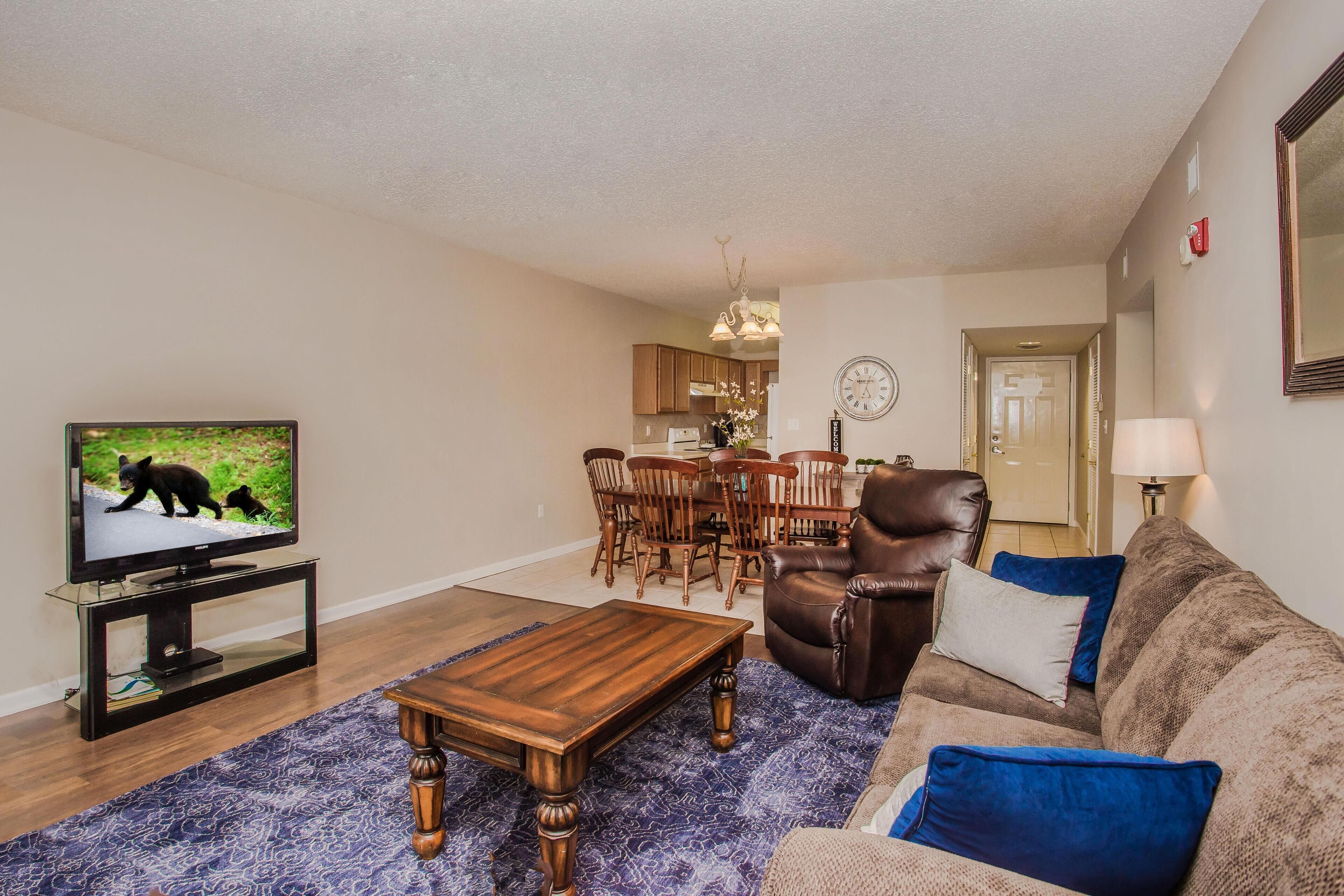 A living room with a television, a wooden coffee table, a brown leather sofa, and a dining area with wooden chairs and a table.