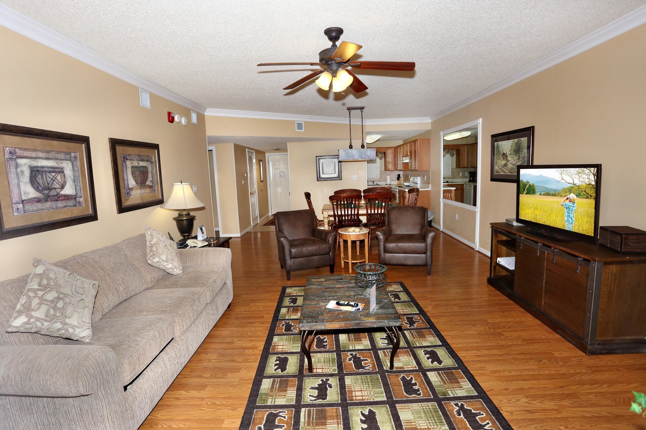 A living room with a sofa, coffee table, and TV. There is a ceiling fan, a dining area with chairs, and a kitchen in the background.
