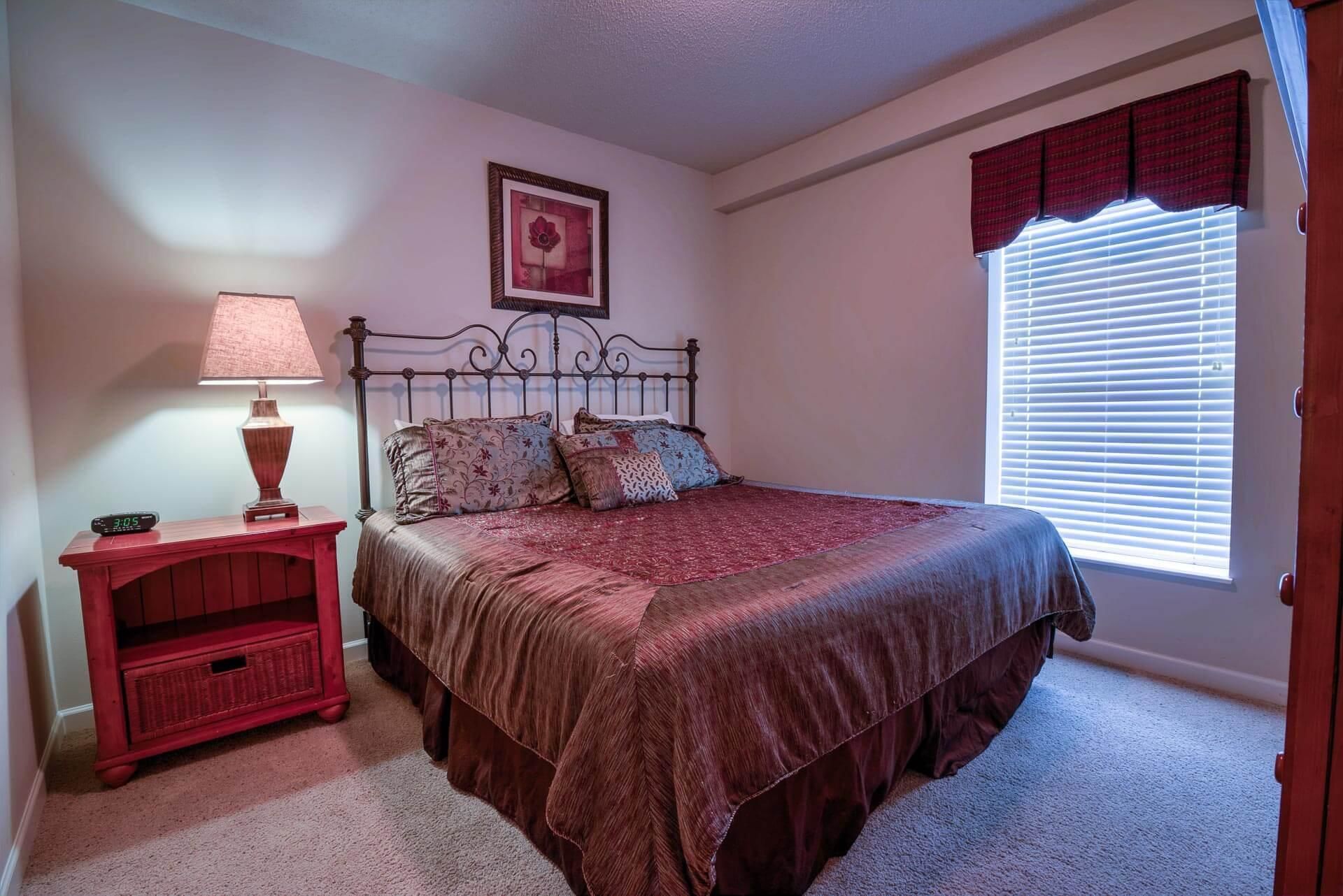 A bedroom with a bed, a nightstand, a lamp, a clock, a window with blinds, and a framed picture on the wall.