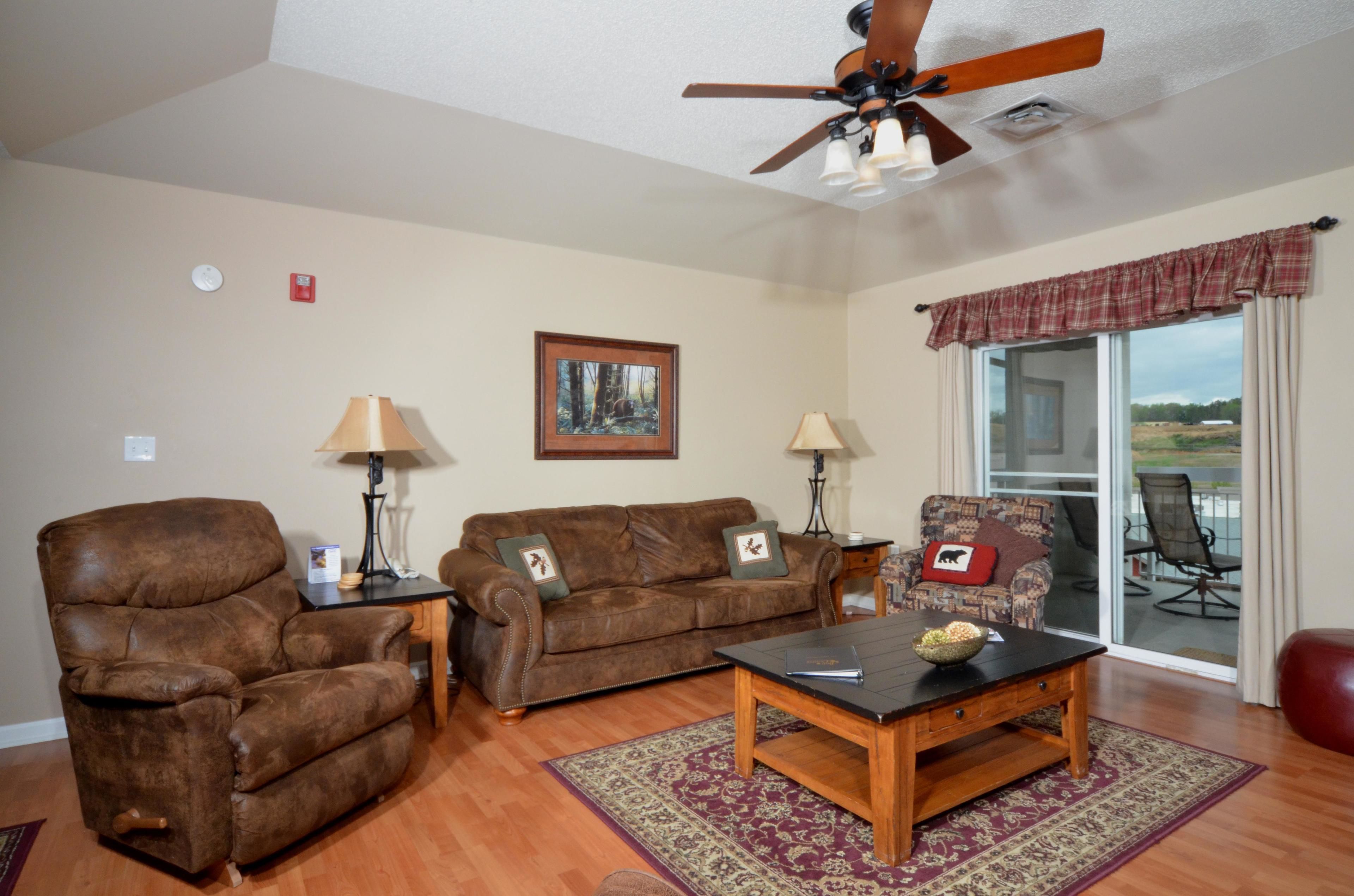 A living room with a brown leather recliner, a brown sofa, a wooden coffee table, and a patterned rug. There is a ceiling fan, a framed picture on the wall, and a view through a glass door.