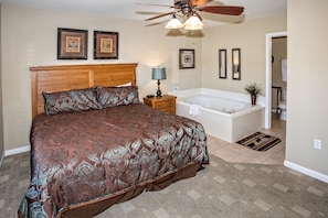 A bedroom with a large bed, a wooden headboard, a nightstand, a lamp, a ceiling fan, and a bathroom with a bathtub and a shower.