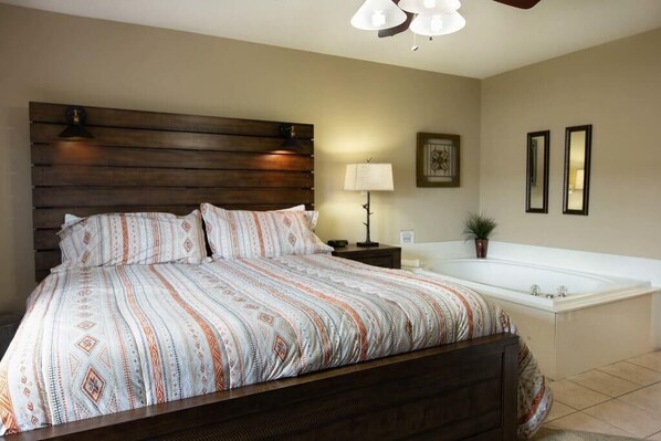 A bedroom with a wooden headboard, a large bed with patterned bedding, a nightstand, a lamp, a framed picture, and a white bathtub.