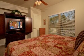 Une chambre à coucher avec un grand lit, un téléviseur posé sur un meuble en bois, une fenêtre avec des stores et un ventilateur de plafond.