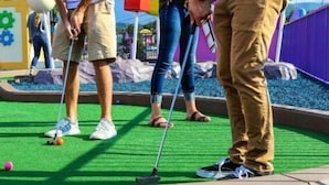 Un campo de minigolf con superficie de juego verde, valla púrpura y un pequeño grupo de personas.