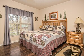 A bedroom with a wooden bed, a nightstand, a lamp, a window with curtains, and a wall with a picture and two small trees.