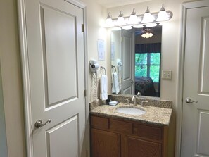 A bathroom with a sink, vanity, and a mirror. There are hair dryers mounted on the wall, and a window with curtains.