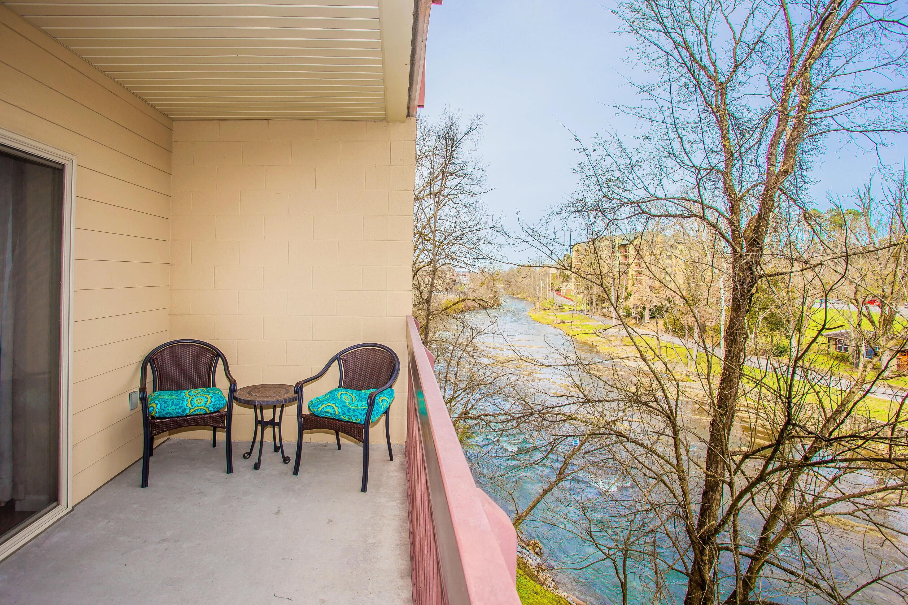 A balcony with two wicker chairs and a small table, overlooking a river and trees.