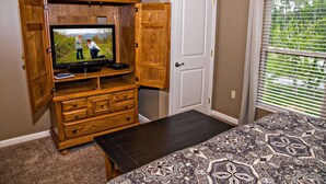 A bedroom with a bed, a wooden entertainment center with a TV, a door, and a window with blinds. - Cedar Lodge 302 (Pigeon Forge)