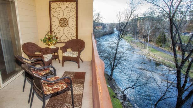 Een balkon met rieten stoelen en een tafel, met uitzicht op een rivier en gebouwen en bomen.