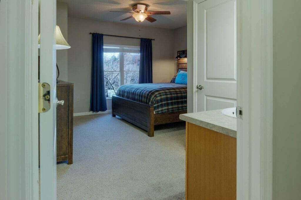 Une chambre à coucher avec un lit, un ventilateur de plafond, une fenêtre avec des rideaux, une table de chevet et une porte.