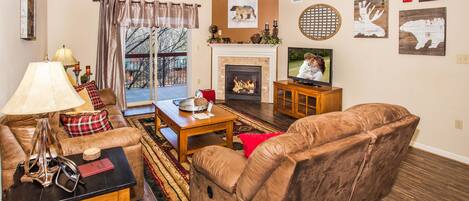 Una sala de estar con sofá, mesa de centro y chimenea. Hay un televisor sobre un mueble bajo y un ventilador de techo.