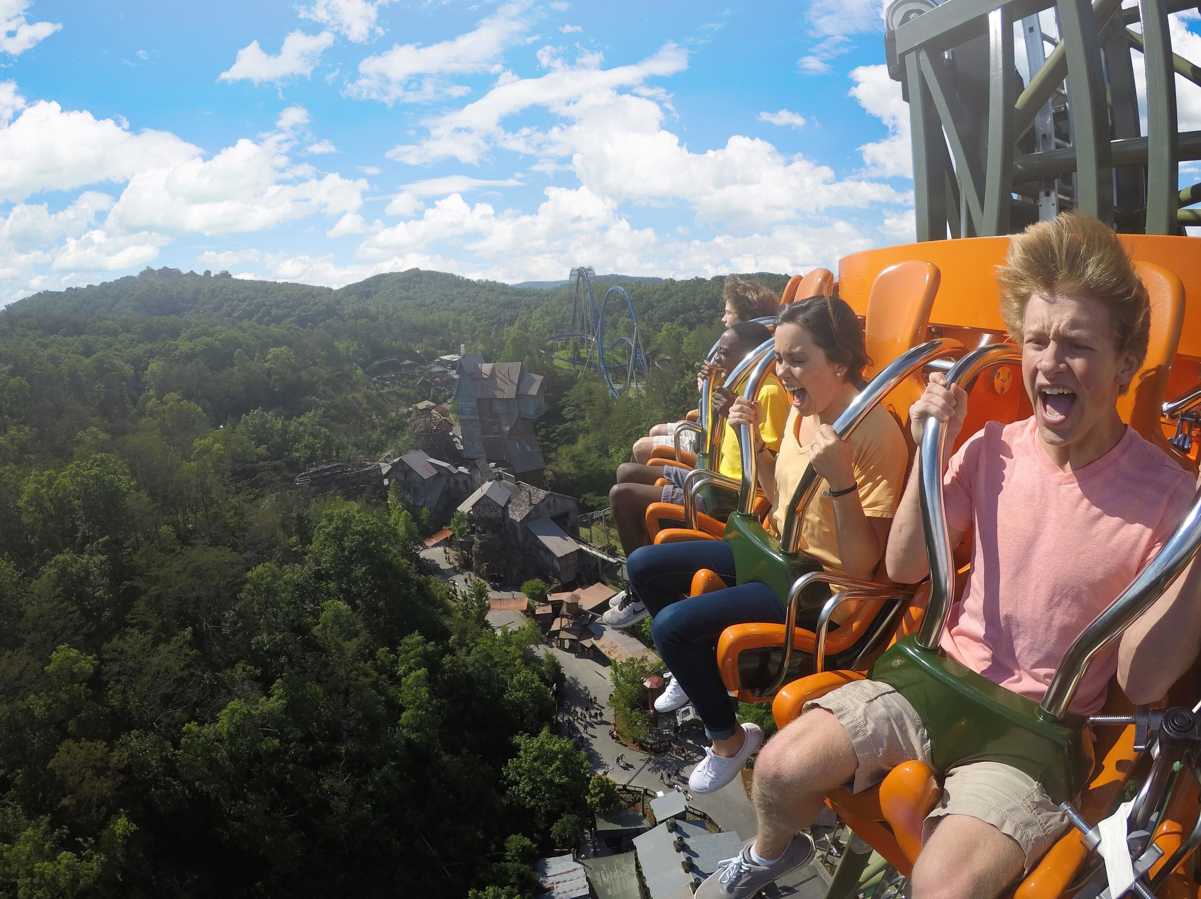 A roller coaster with passengers seated in rows, surrounded by lush greenery and a clear sky.