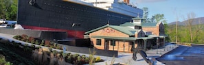 A large cruise ship docked next to a brick building with a green roof, possibly a restaurant or bar, and a parking area with a water feature. - Cedar Lodge 504 (Pigeon Forge)