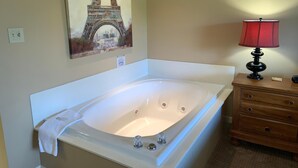 A bathroom with a large bathtub, a wooden nightstand with a lamp, and a painting of the Eiffel Tower.