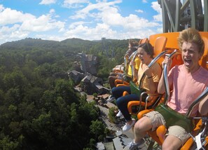 A roller coaster with passengers seated in orange cars, surrounded by a forested landscape and a clear sky. - Cedar Lodge 605 (Pigeon Forge)