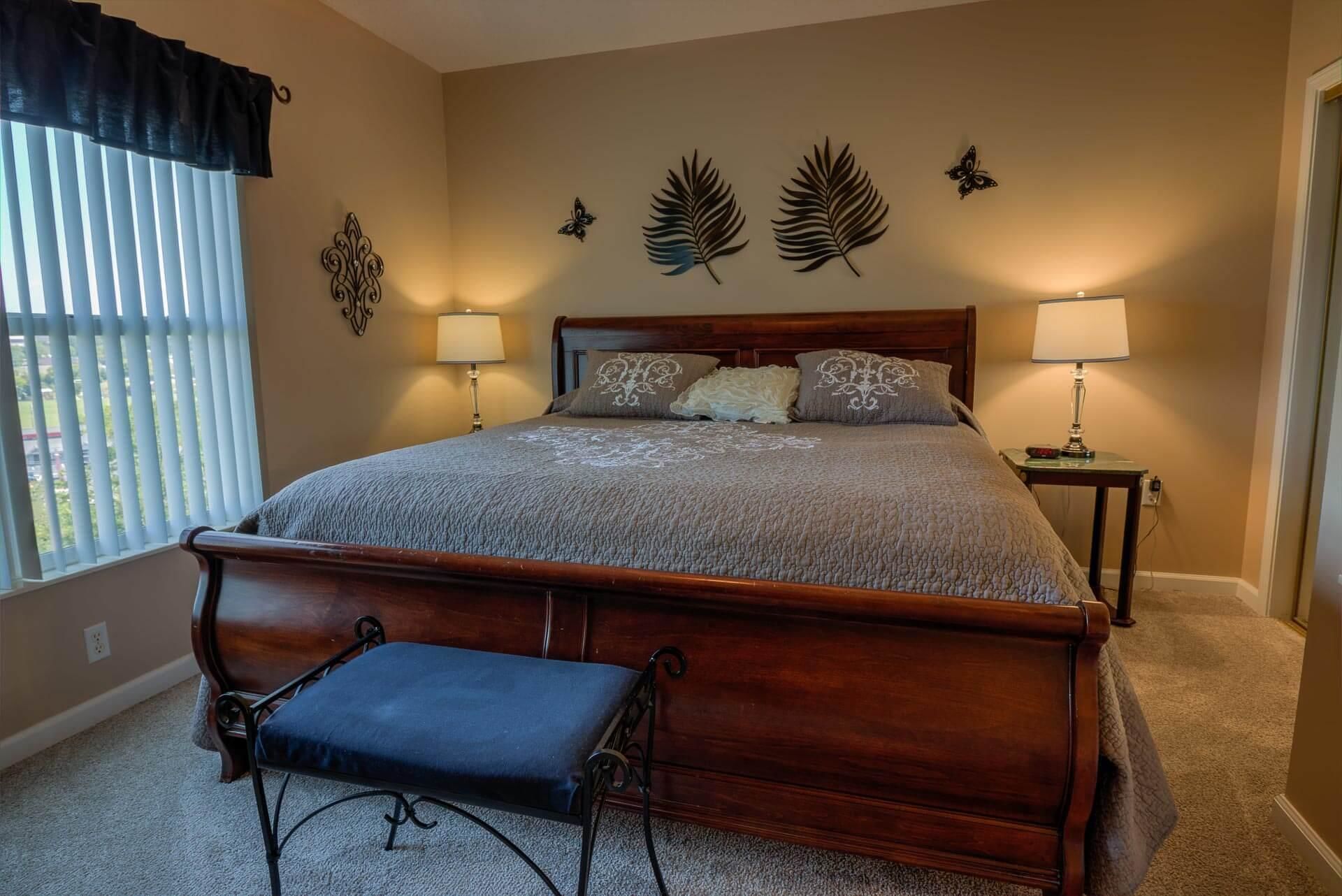 Un dormitorio con cama de madera, dos lámparas de noche, decoración en la pared, un banco y una ventana con persianas.
