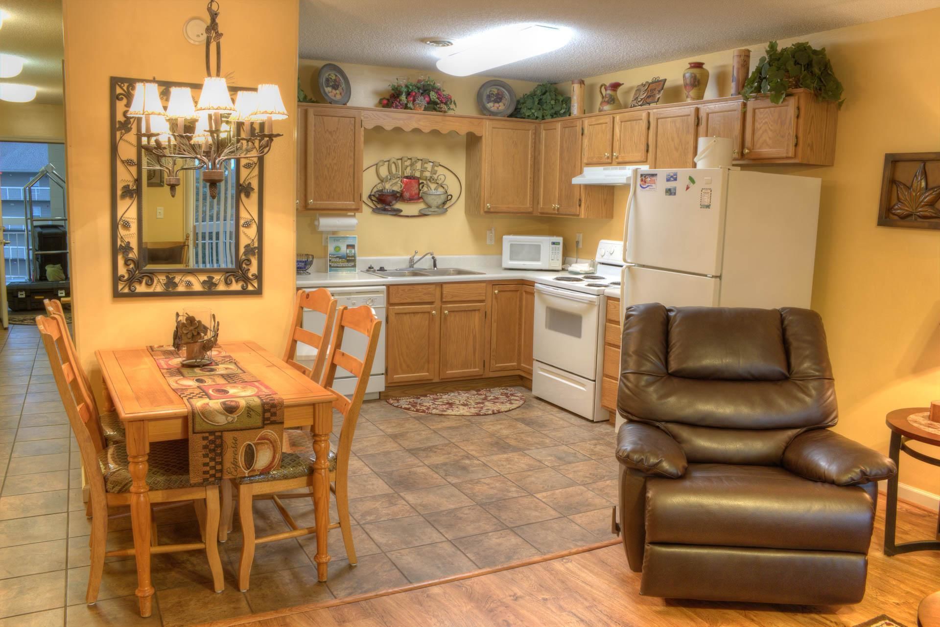 A compact kitchen with wooden cabinets, a dining table with chairs, a leather armchair, and a refrigerator.