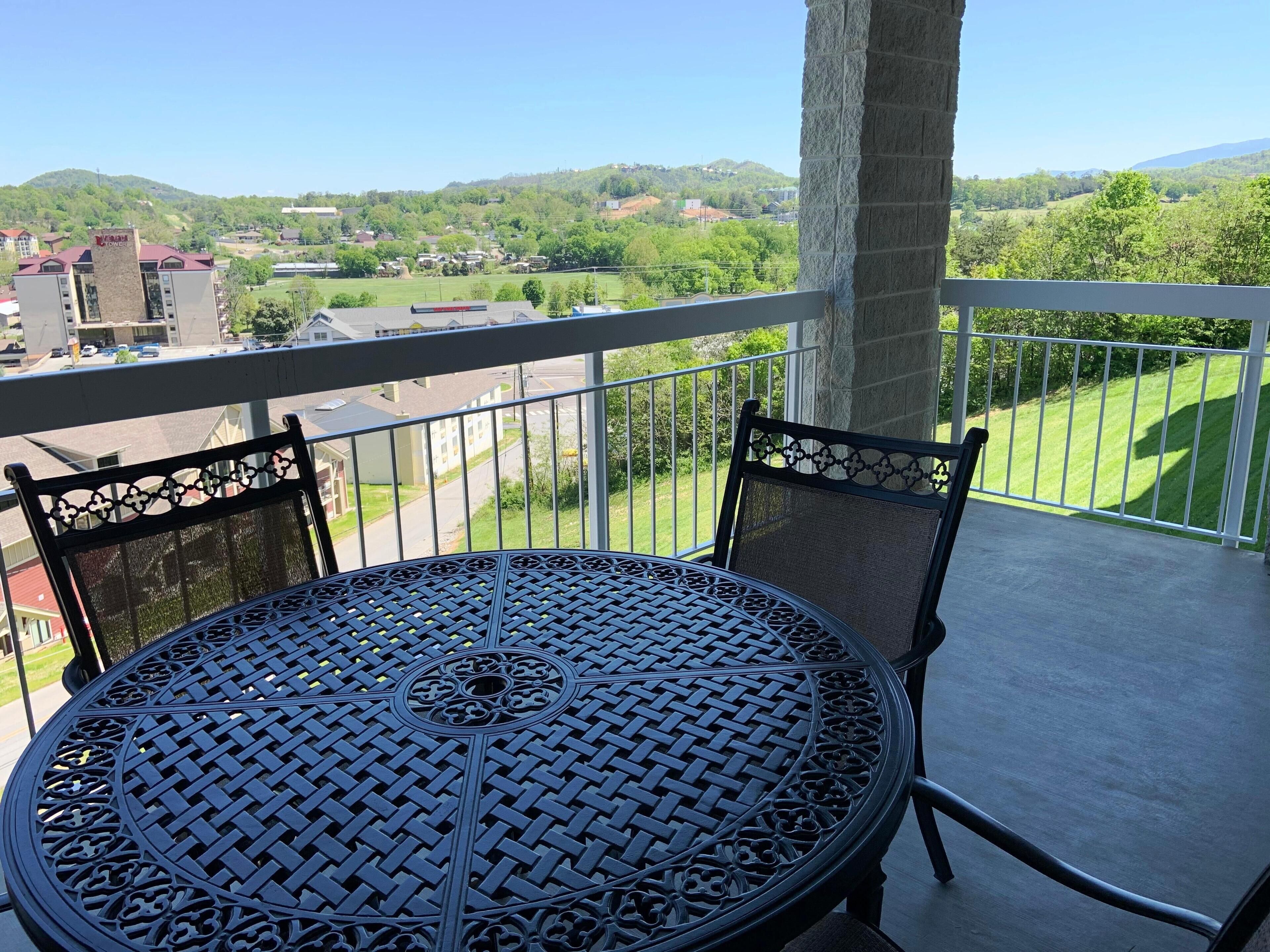Ein Balkon mit einem runden Metalltisch und zwei Stühlen, mit Blick auf eine grüne Landschaft und Gebäude in der Ferne.