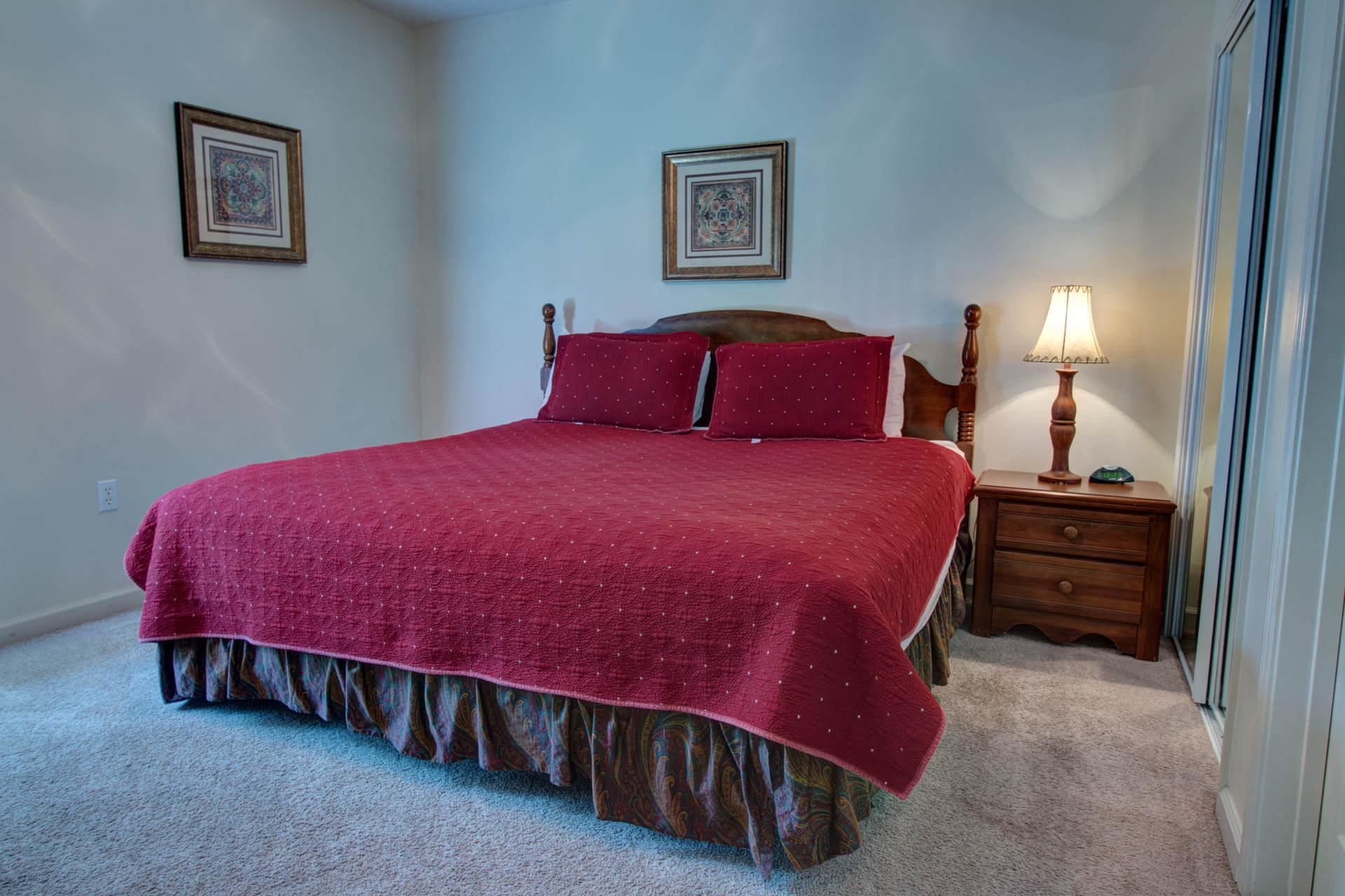 A neatly made bed with a red bedspread and matching pillows, a wooden headboard, a nightstand with a lamp, and two framed pictures on the wall.