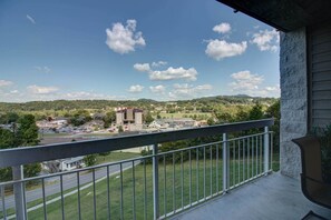 A balcony with a metal railing overlooking a town with a mix of residential and commercial buildings, a parking lot, and green hills in the distance. - Whispering Pines 214 (Pigeon Forge)