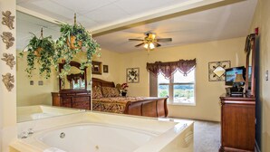 A bedroom with a large white bathtub, a ceiling fan, a bed with a floral bedspread, a dresser, and a television. - Whispering Pines 212 (Pigeon Forge)